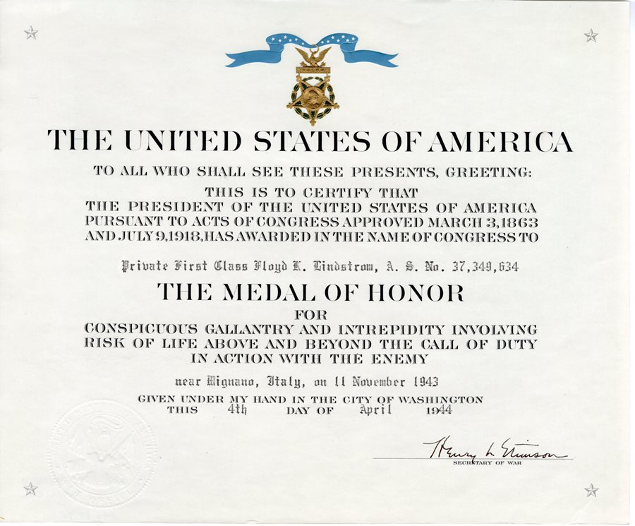 USA Medal of Honor Certificate that is white with an image of the medal shaped a little like a bell with a blue ribbon tied to the top of the medal. The script relates to Floyd's acts of valor on the hills near Mignano, Italy on November 11, 1943, Lindstrom was nominated for the Medal of Honor.