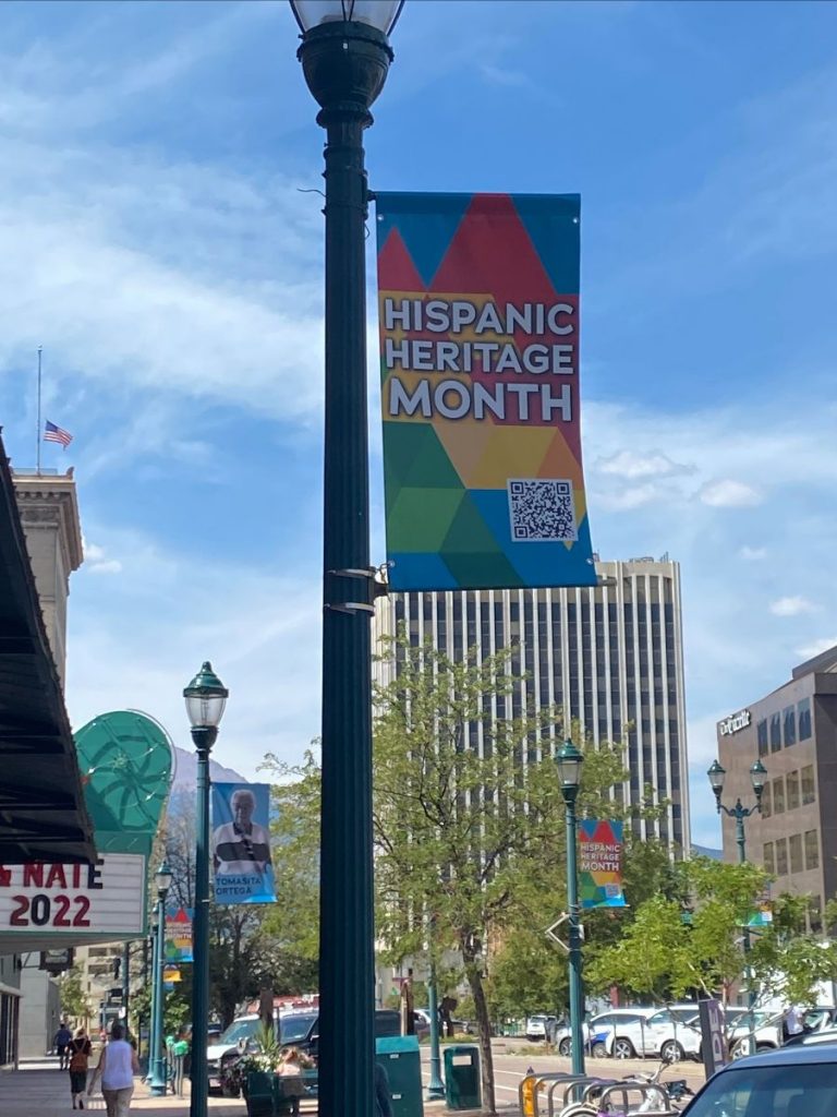 A colorful banner mounted on a light post reads "Hispanic Heritage Month". In the background is Pikes Peak Ave. in Colorado Springs, Colorado.