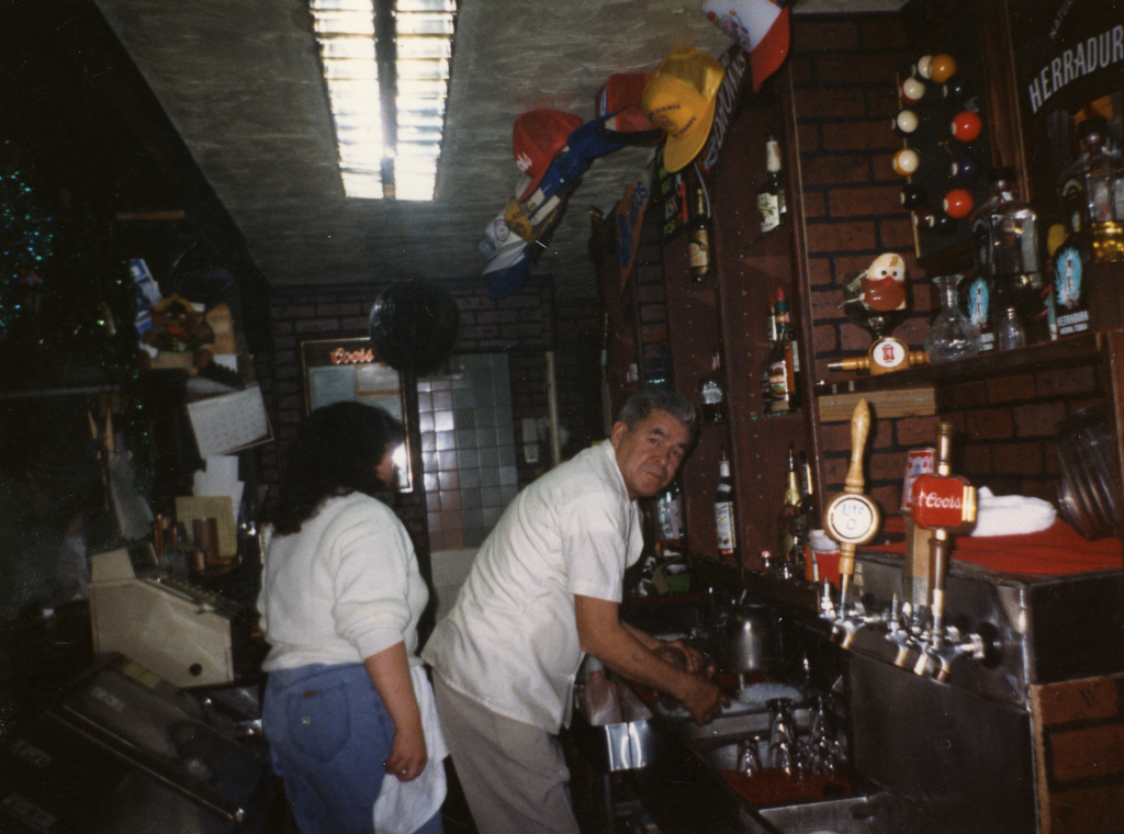 Victor Ornelas looks toward the camera while leaning over a sink at a bar. A woman passes by behind him looking away from the camera. Liquor bottles can be seen on shelves.