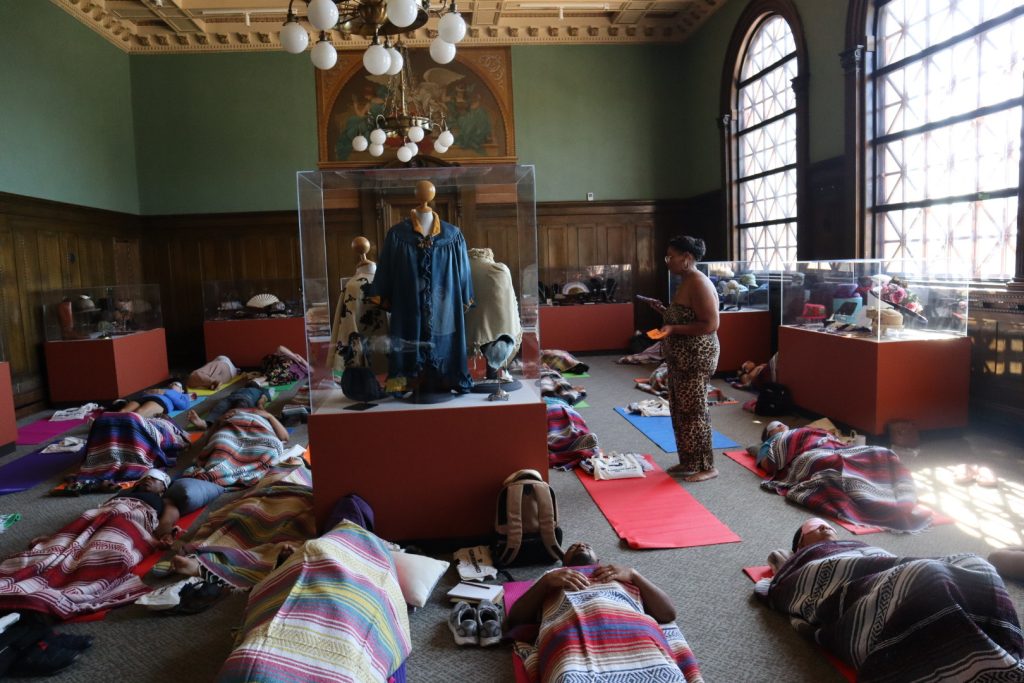 Program participants lay on the floor of the "Accessorize It!" exhibit on top of colorful yoga mats while the program presenter, Ashley Cornelius, walks around them.