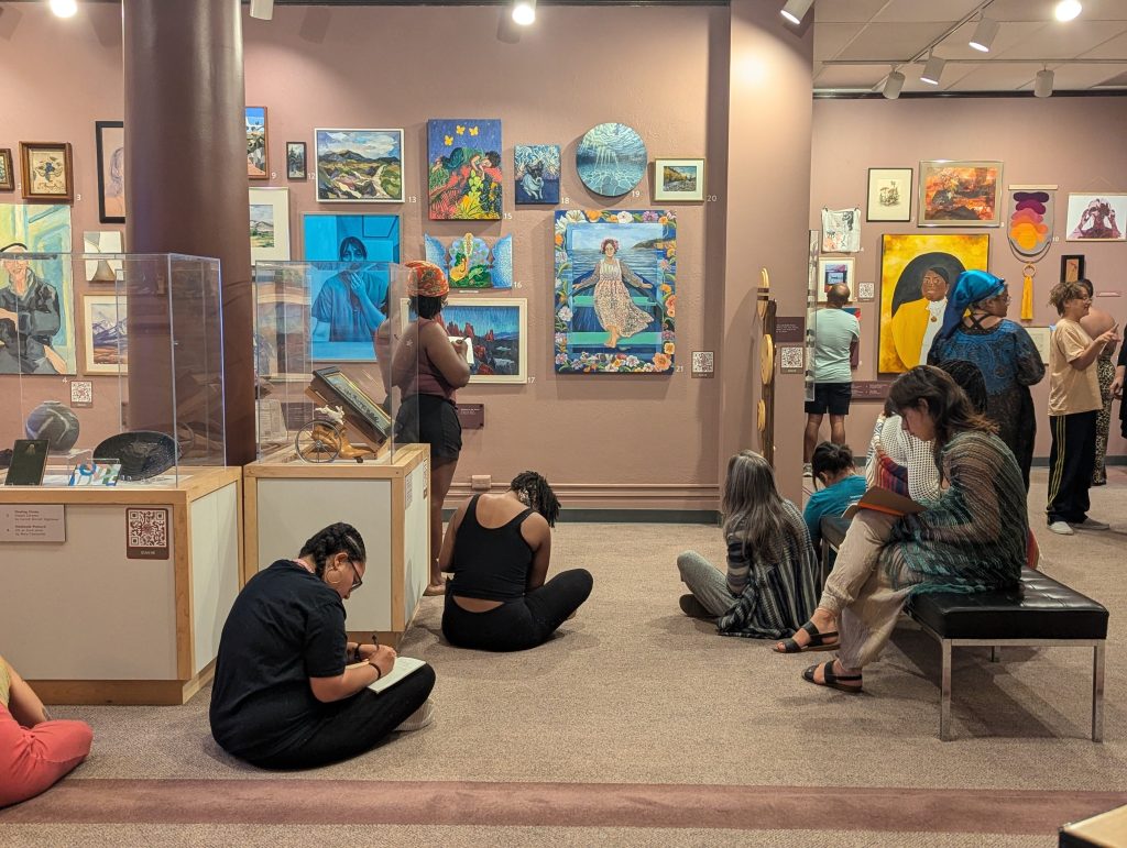 Program participants sit on the floor and a bench and walk around the "50% of the Story" exhibit. They are writing in notebooks and looking at the art on the walls.