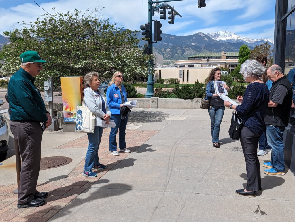 A group of people are gathered outside on a downtown Colorado Springs street. One person is showing historic photographs to the others. They are practicing leading walking tours for the Colorado Springs Pioneers Museum.