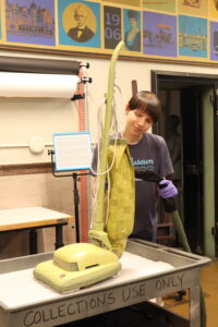 Brayan Montes De Oca uses the Nilfisk Vacuum to remove dust from a 1970s Hoover Convertible Vacuum Model 707 cleaner. To protect the object during this, pantyhose is used to cover the vacuum’s nozzle of the vacuum.